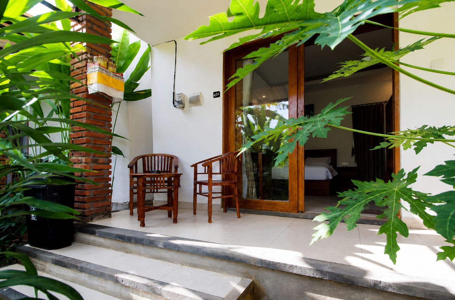 A room with a bed, a wooden chair, a potted plant, and a brick wall.