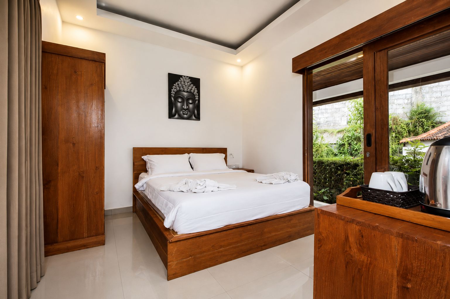 A bedroom with a wooden bed, white bedding, a Buddha statue on the wall, and a view of greenery outside.