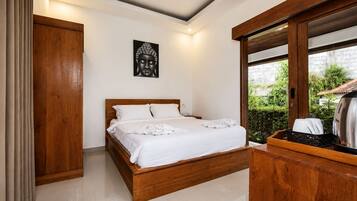 A bedroom with a wooden bed, white bedding, a Buddha statue on the wall, and a view of greenery outside.