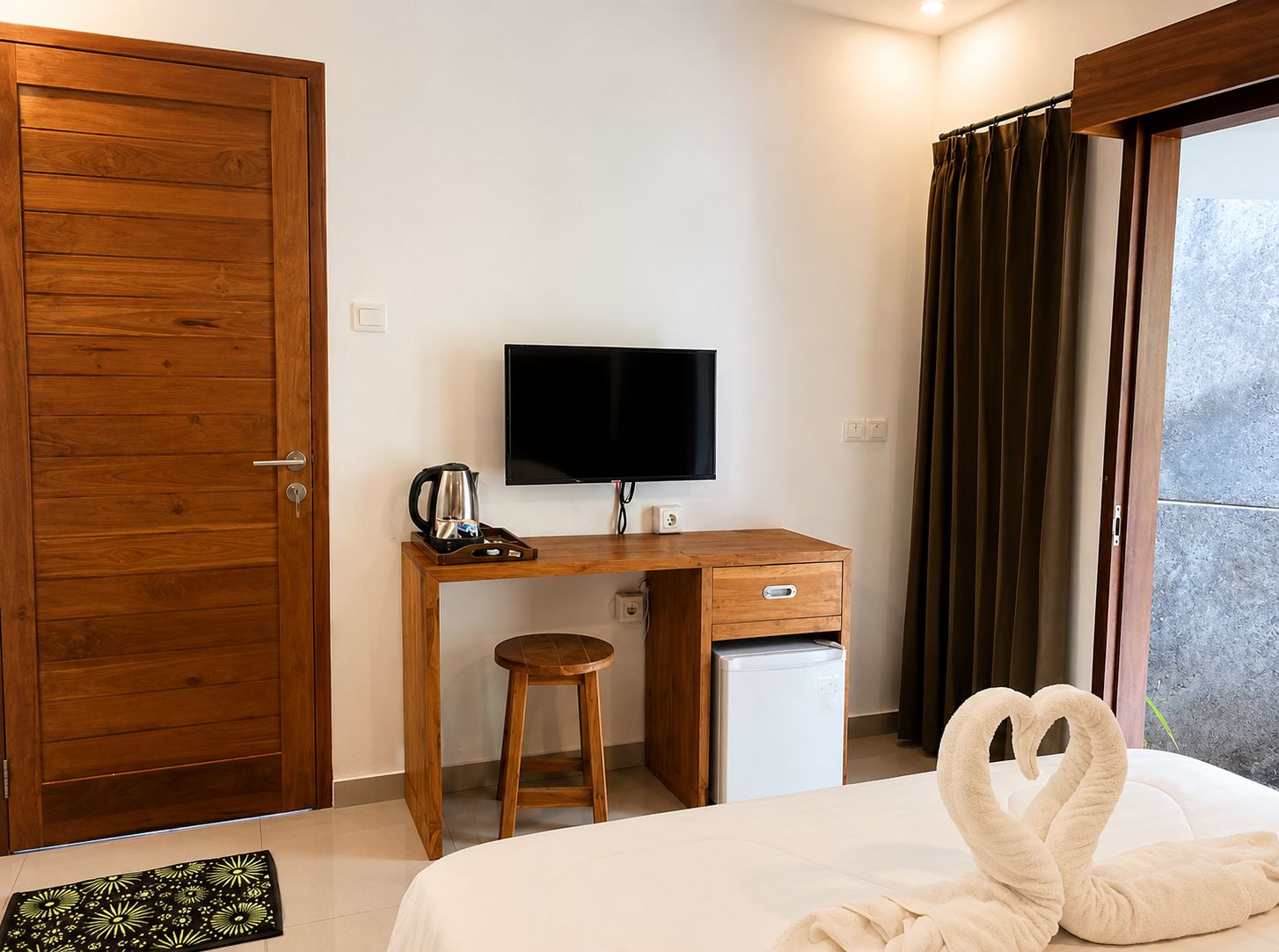 A hotel room with a wooden desk, a television, a small refrigerator, a bed with a swan-shaped pillow, and a window with curtains.