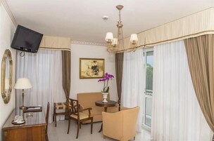 Una camera d'hotel con televisione, un divano, una poltrona, un tavolino con un vaso di fiori e una finestra con tende.