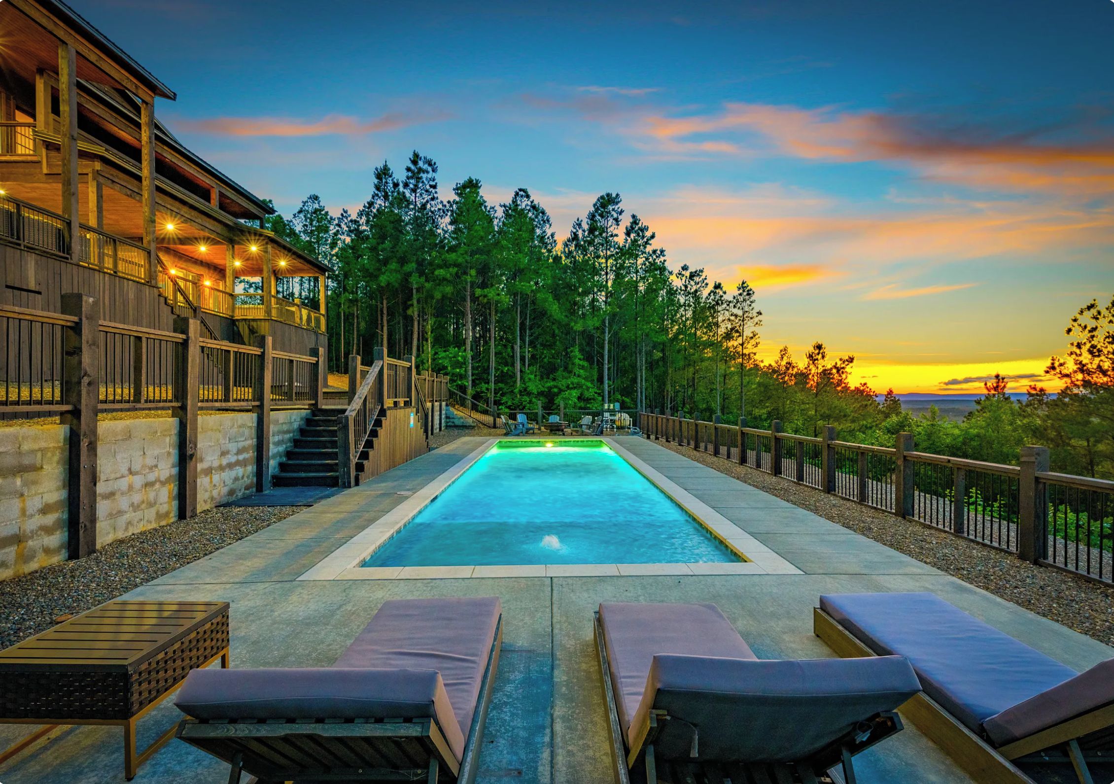 A swimming pool with lounge chairs, a wooden deck, and a view of trees and a sunset.