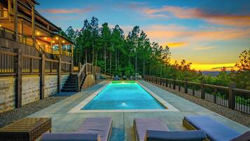 A swimming pool with lounge chairs, a wooden deck, and a view of trees and a sunset.