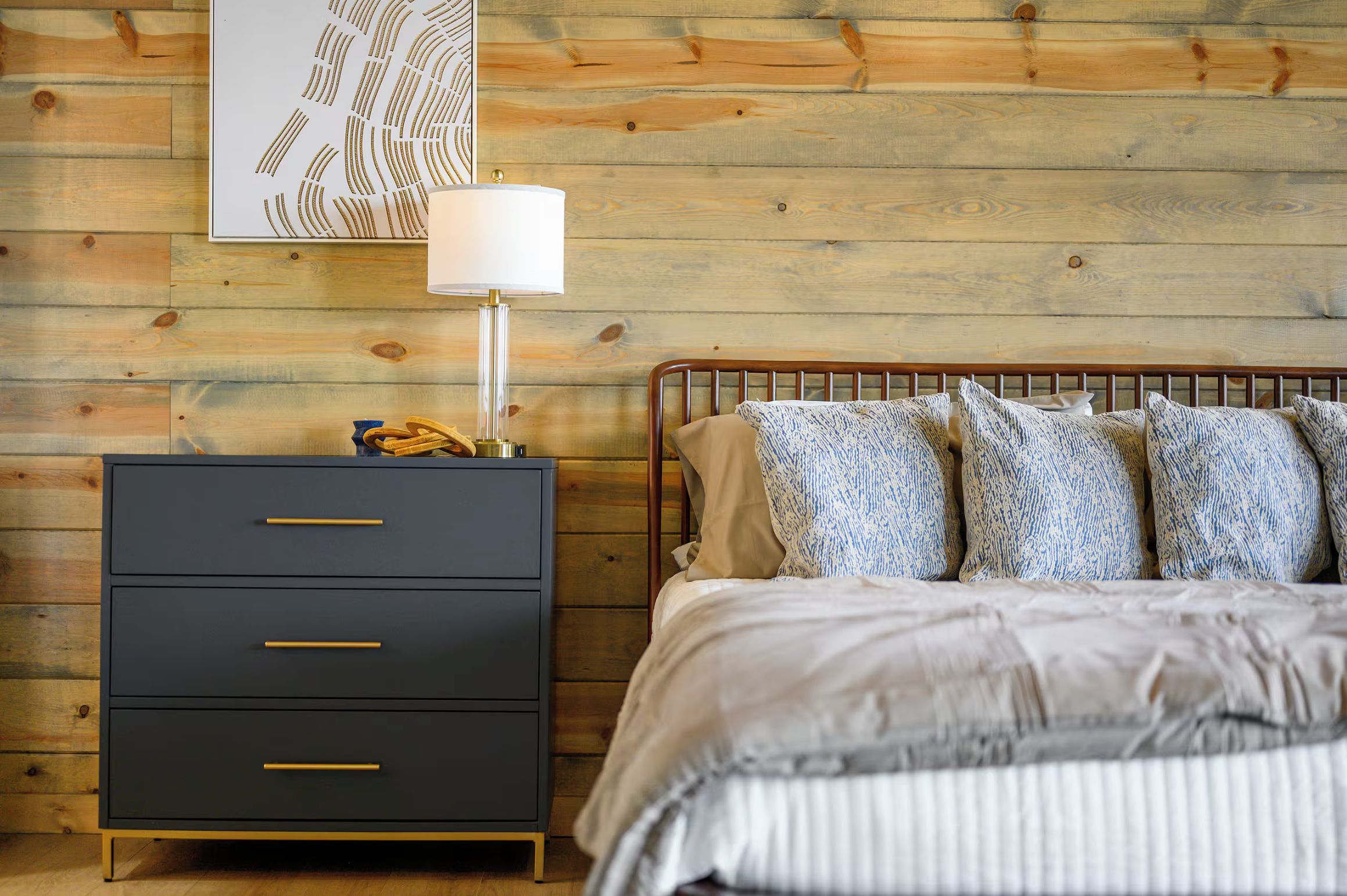 Quarto com parede de madeira, cama com almofadas estampadas, mesa de cabeceira com abajur e cômoda escura com puxadores dourados.