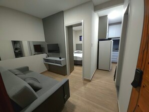 A modern living room with a grey sofa, a TV mounted on the wall, and a kitchen area visible through an open door. - Flat cidade do leite (Nossa Senhora da Glória)