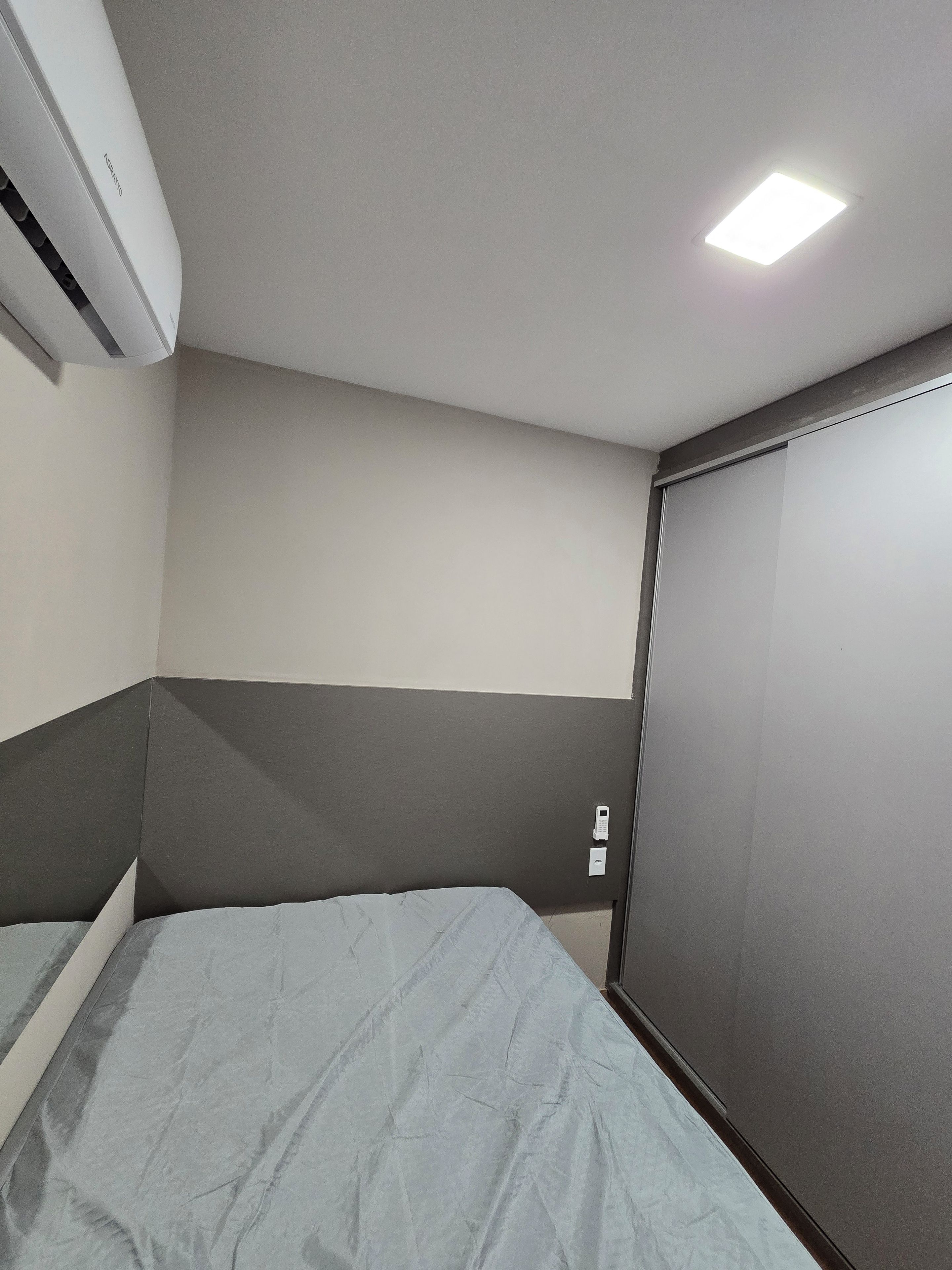 Quarto com cama, ar condicionado na parede, luminária no teto e uma porta.