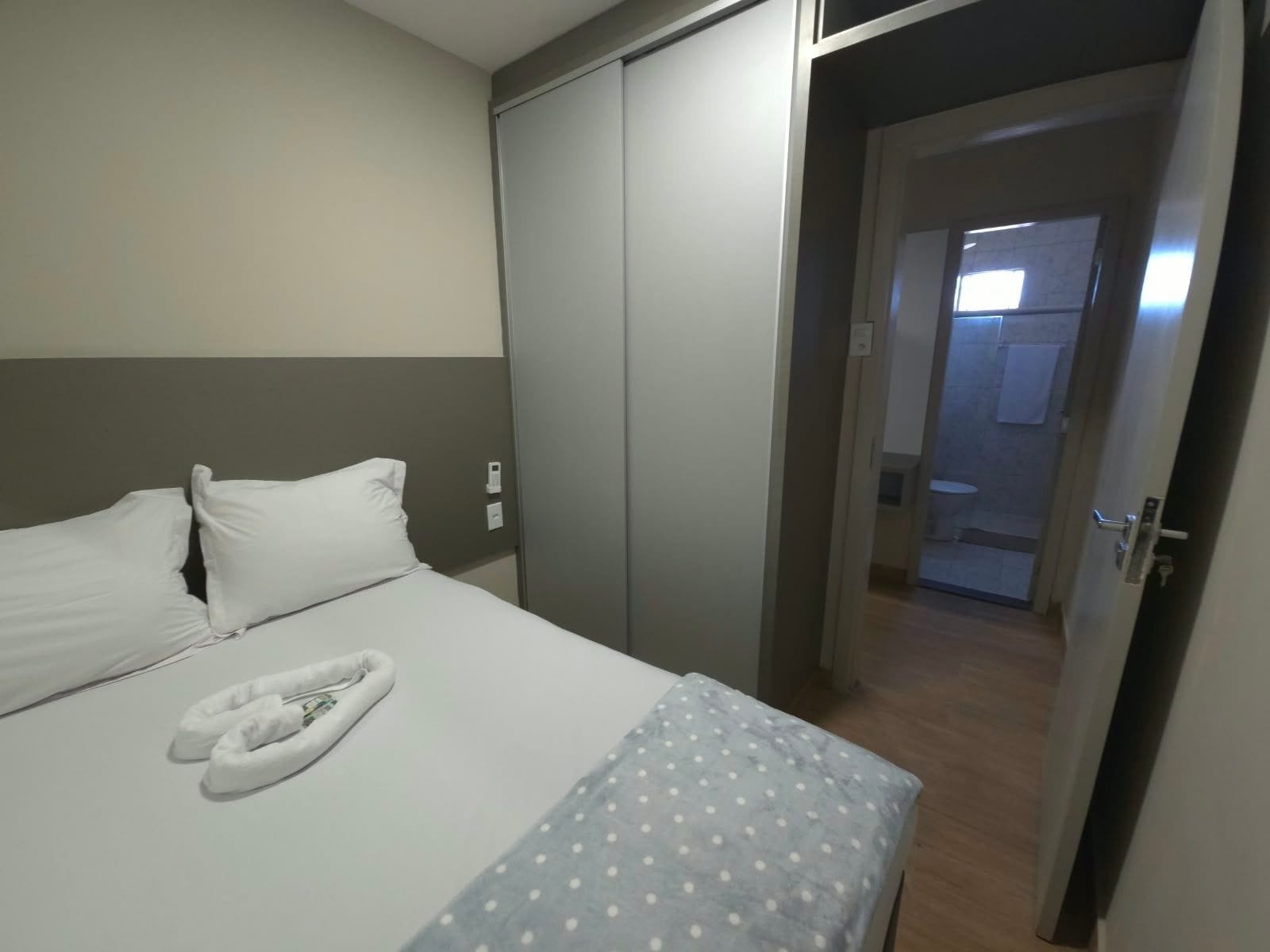 A hotel room with a bed, pillows, a folded towel, and a door leading to a bathroom.