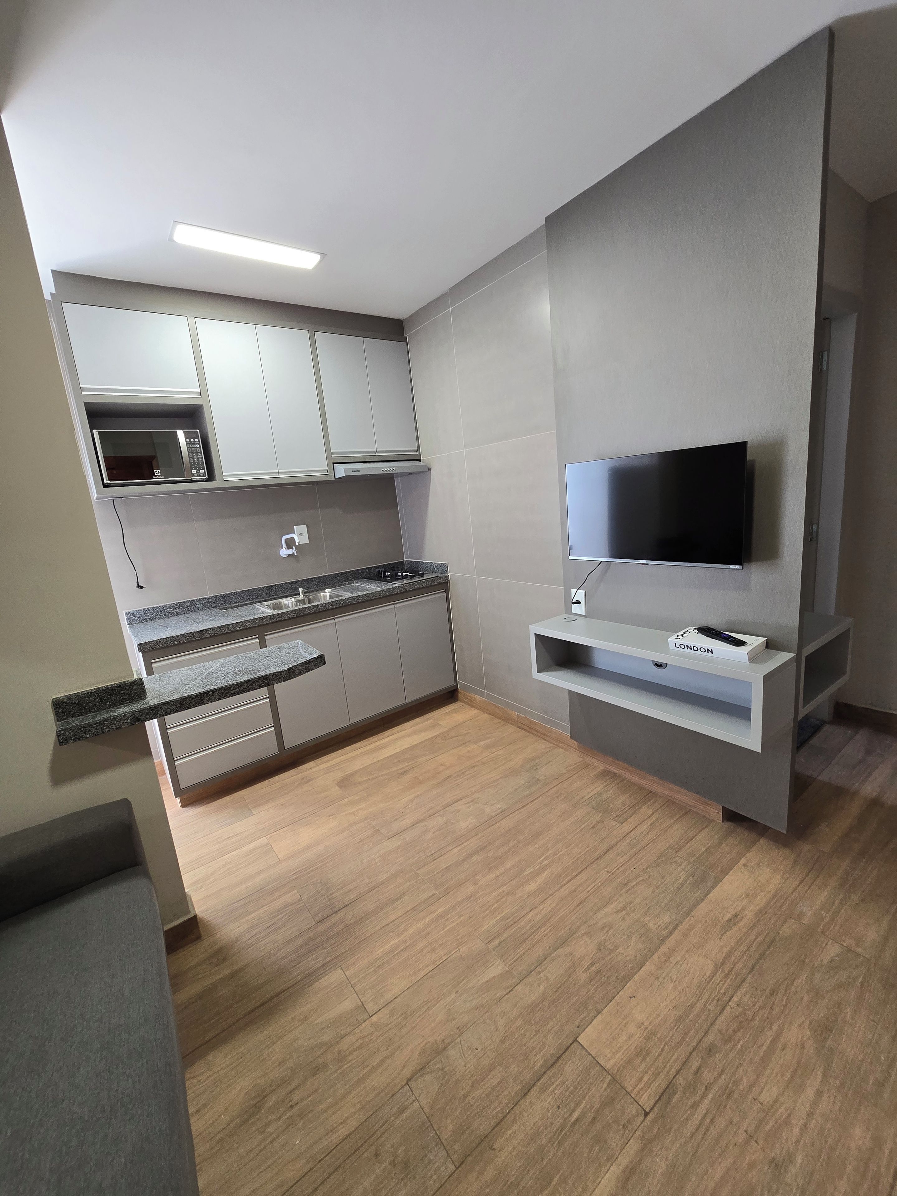 A modern kitchen with grey cabinets, a built-in microwave, a flat-screen TV, and a wooden floor.
