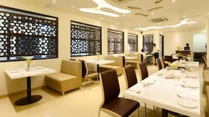 Ein Restaurant mit mehreren Tischen und Sitzecken, manche mit weißen Tischdecken, andere ohne. Dekorative Fensterläden und ein modernes Interieur mit einer neutralen Farbgestaltung.
