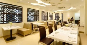 A restaurant with multiple tables and booths, some with white tablecloths and others with no tablecloths. There are decorative screens on the windows, and the interior has a modern design with a neutral color palette. - Hotel Madhushrie (Etmadpur)