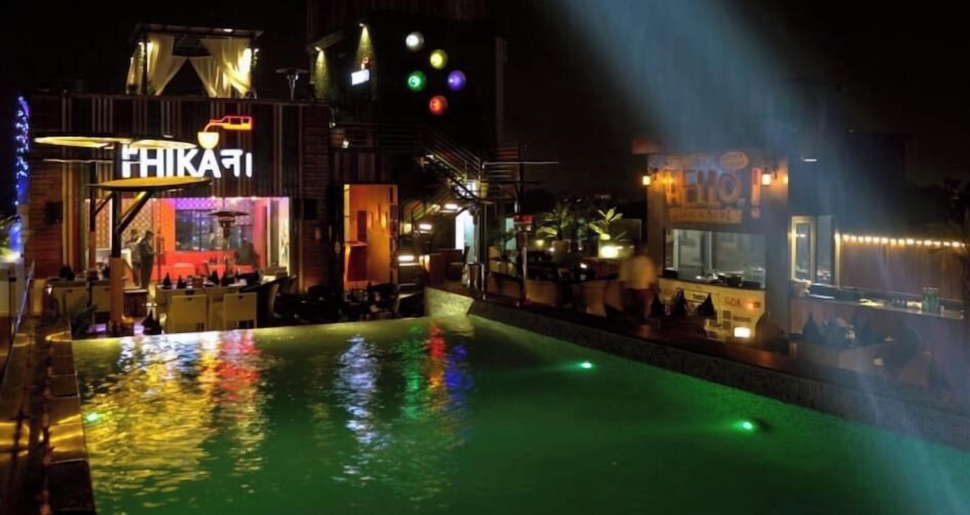 Un área de piscina al aire libre con un bar y asientos, rodeada de edificios con letreros iluminados.