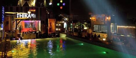 An outdoor pool area with a bar and seating, surrounded by buildings with illuminated signs.