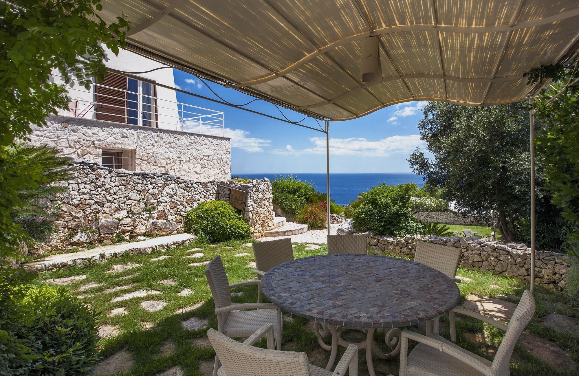 A patio with a stone table and wicker chairs, overlooking a stone building and the sea.