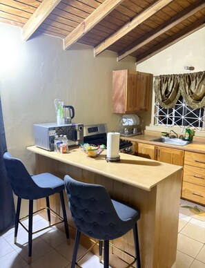 A kitchen with a wooden island, two bar stools, a coffee maker, and a window with curtains. - 🌴 New Harbour Haven | Spacious Family Retreat in Gated Community (Old Harbour)