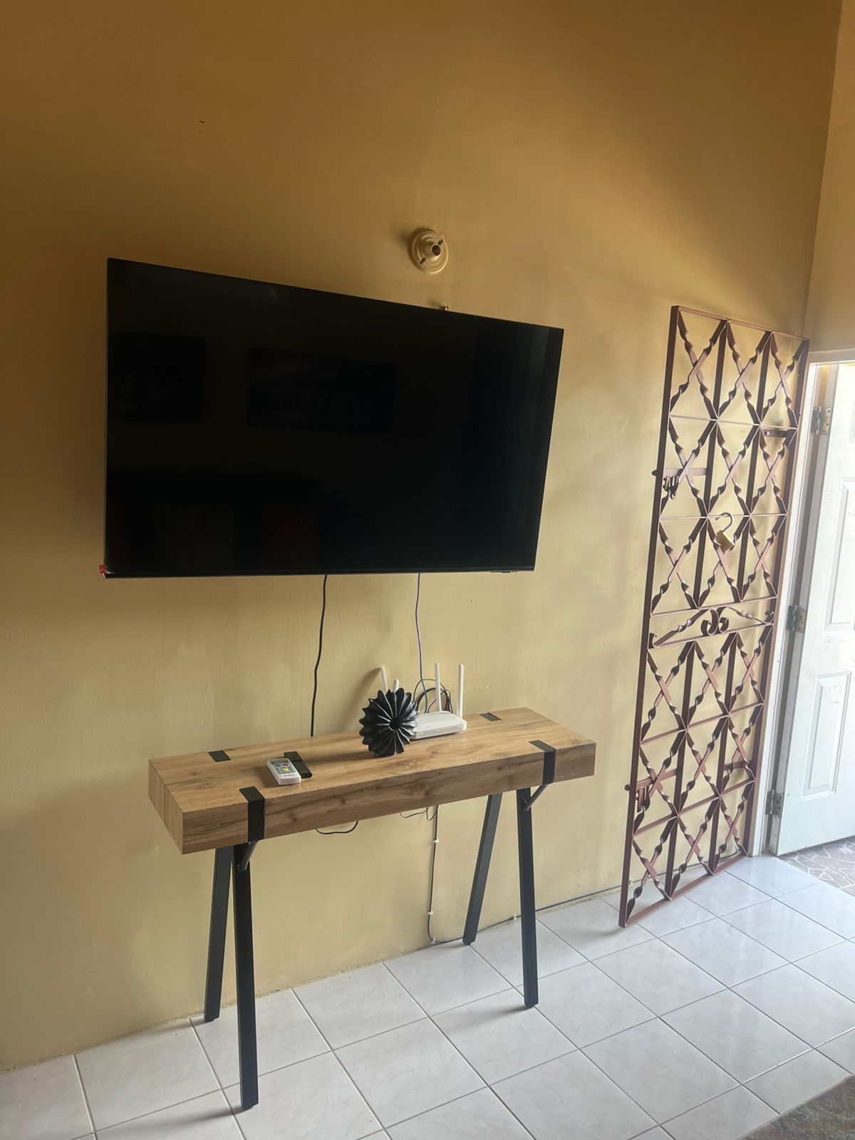 Un televisor de pantalla plana montado en una pared, una consola de madera con un módem y un objeto decorativo, y una partición metálica con diseño.