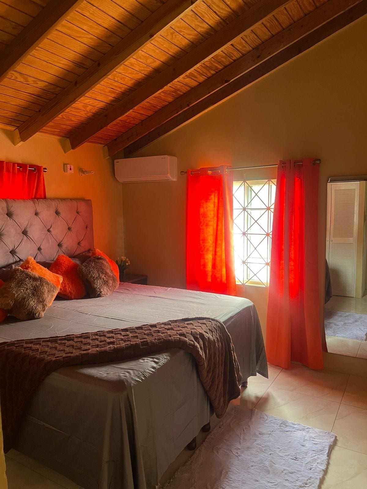 A bedroom with a large bed, a wooden ceiling, and a window with red curtains.
