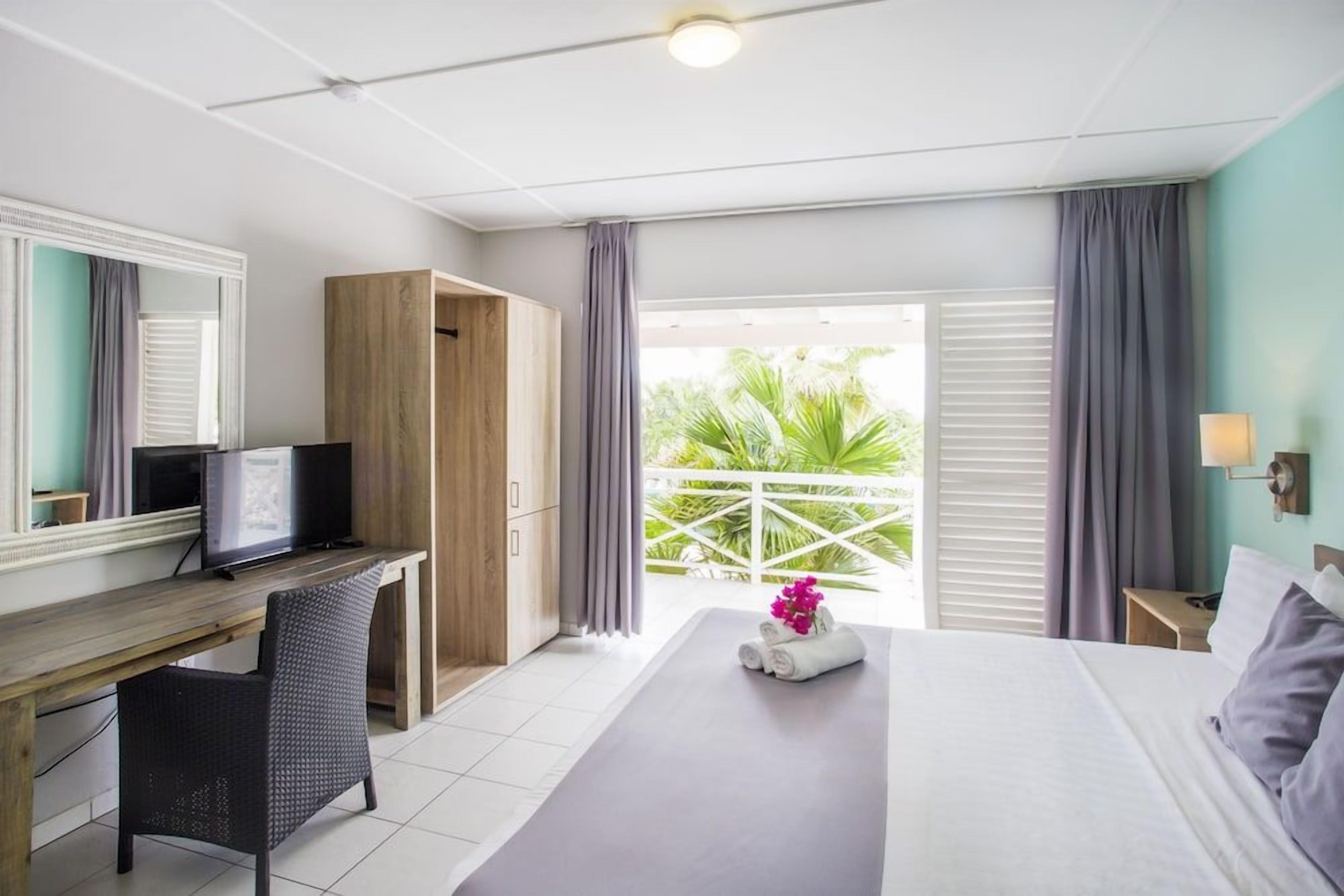 Een hotelkamer met een bed, een bureau met een computer, een stoel, een kledingkast en een balkon met uitzicht op groen.