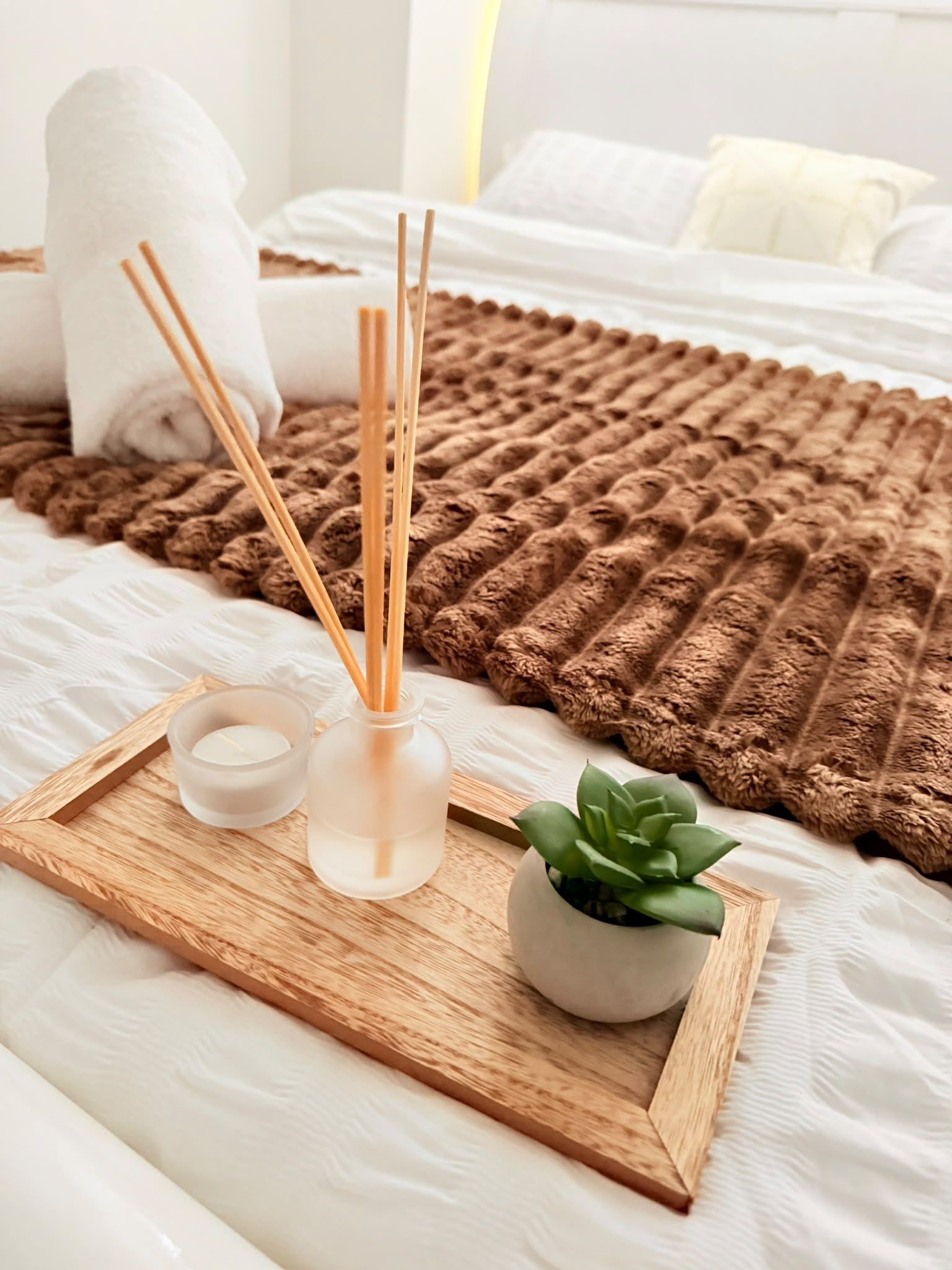 Un lit bien fait, recouvert d’une couverture marron à texture intéressante, avec une petite plante en pot et deux récipients en verre contenant des diffuseurs à bâtonnets, le tout posé sur un plateau en bois.