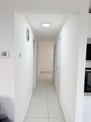 Un corridoio stretto con pareti bianche, porte, pavimento piastrellato e una luce al soffitto.
