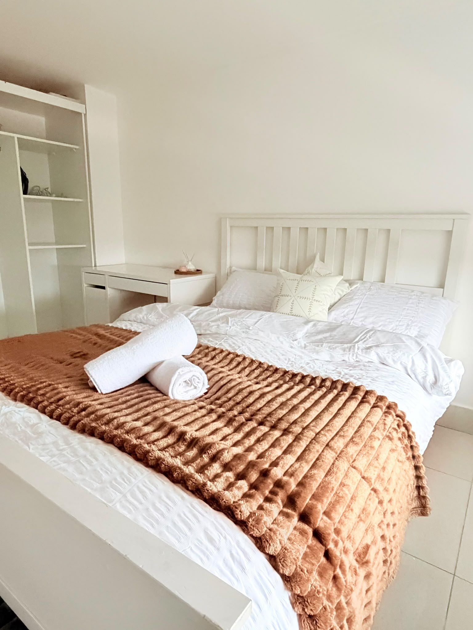 A neatly made bed with a brown blanket and rolled white towels, a white headboard, and a white nightstand with a small item on it.