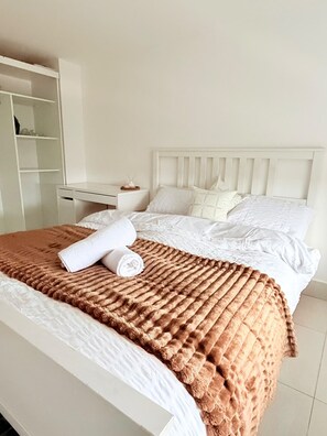 A neatly made bed with a brown blanket and rolled white towels, a white headboard, and a white nightstand with a small item on it. - M C R Skyline View 2-br Apartment w/ Free Parking (Manchester)