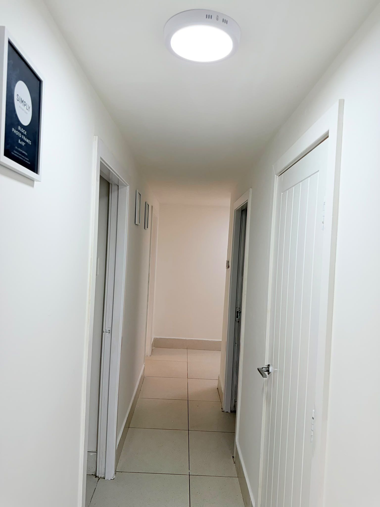 Um corredor estreito com paredes brancas, uma luminária no teto e uma porta à direita.