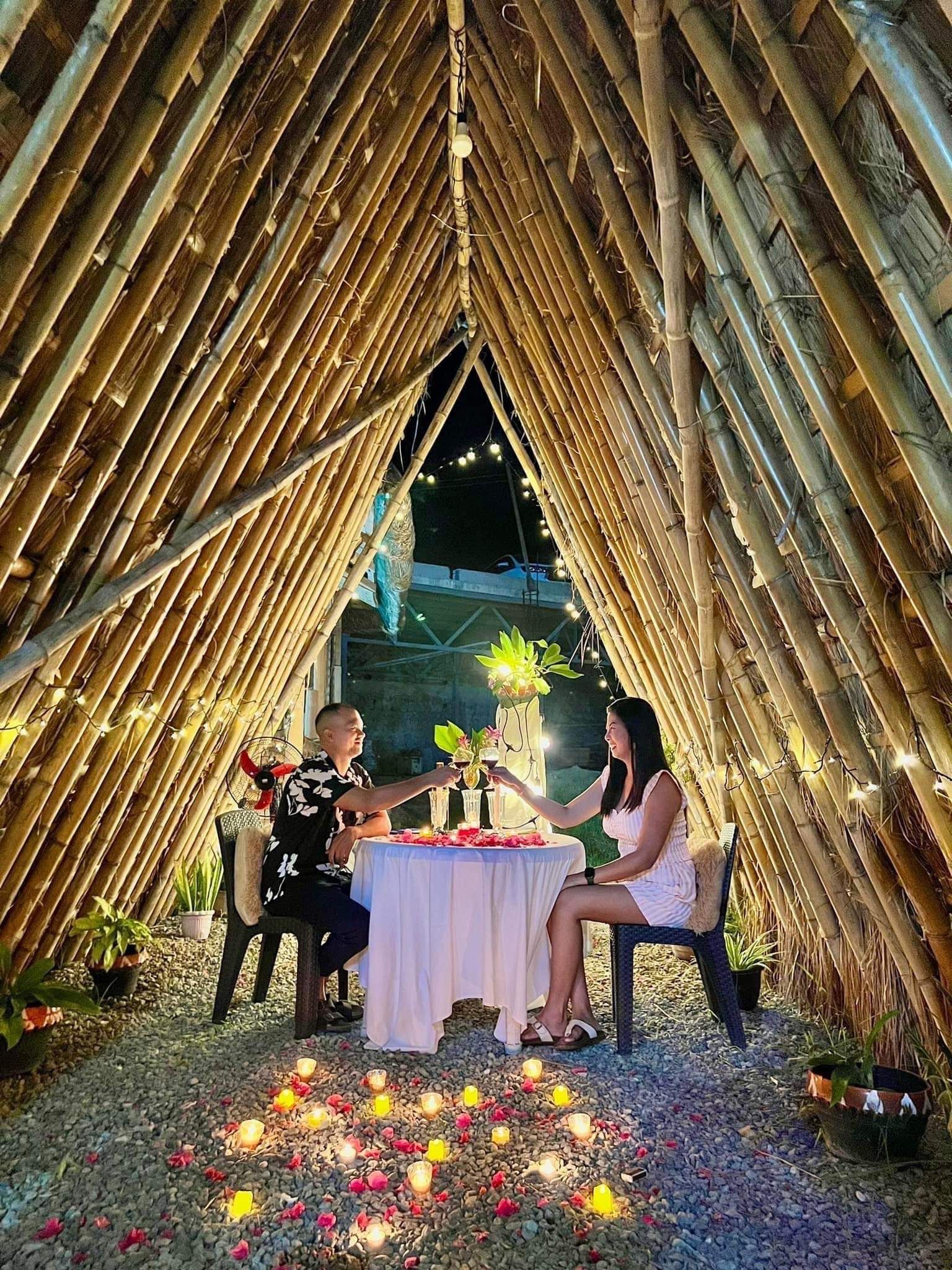 Estrutura de bambu com mesa posta para dois, cercada por pétalas de flores e velas espalhadas.