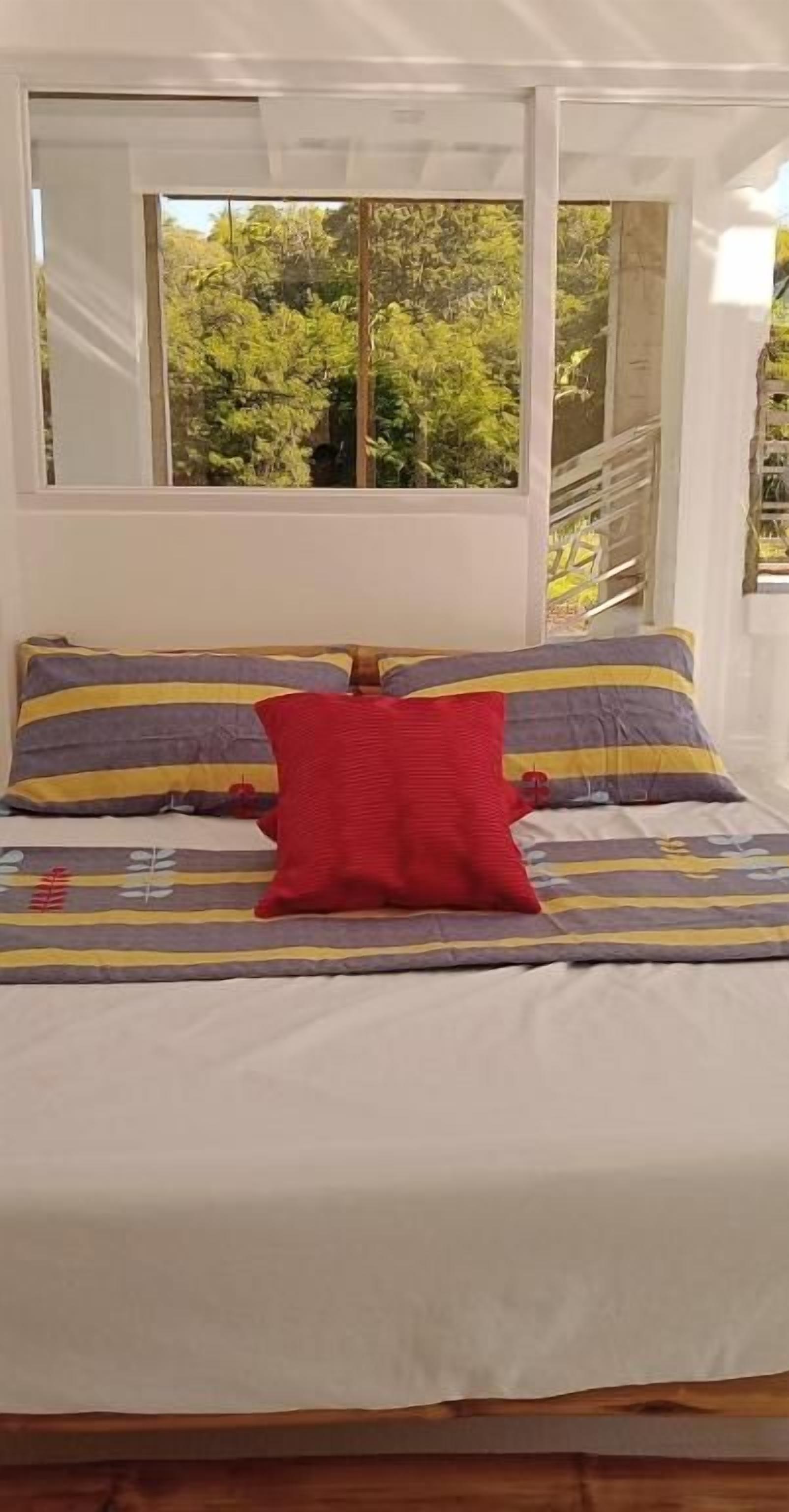 Una cama con un colchón blanco, dos almohadas y un cojín decorativo rojo.
