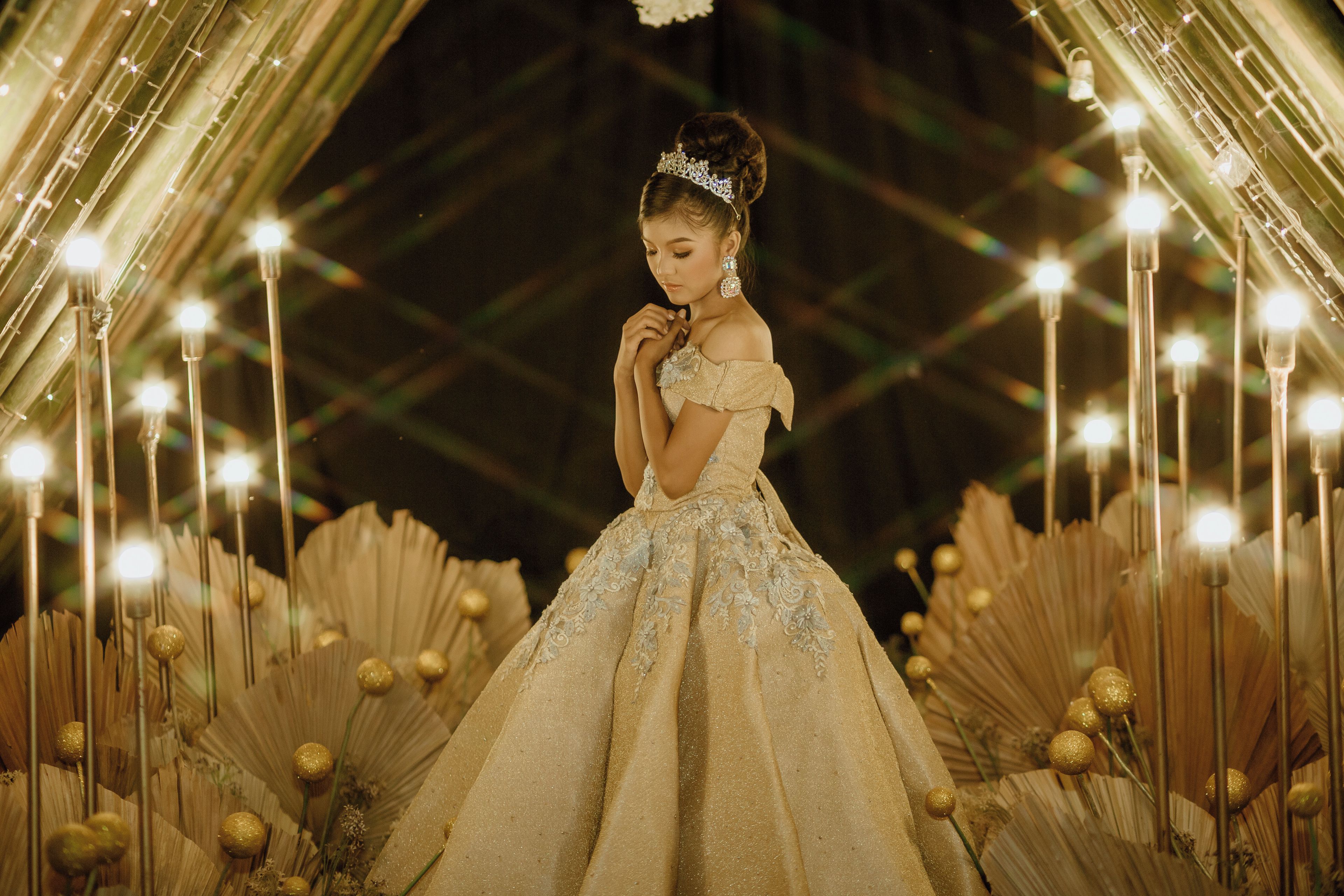 Una persona con un elegante vestido de gala con mangas caídas y una tiara, posando en un entorno ornamentado con iluminación decorativa y arreglos florales.