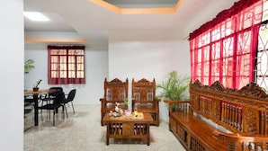 A dining area with a wooden table and chairs, a bench with intricate carvings, and a window with red curtains. - SMyleINN Farm Resort OPC (San Fernando)