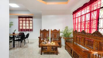 A dining area with a wooden table and chairs, a bench with intricate carvings, and a window with red curtains.