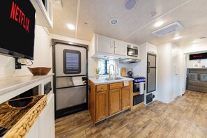 Une cuisine compacte pour camping-car, équipée d’un four à micro-ondes, d’une cuisinière, d’un évier et d’un téléviseur à écran plat fixé au mur.