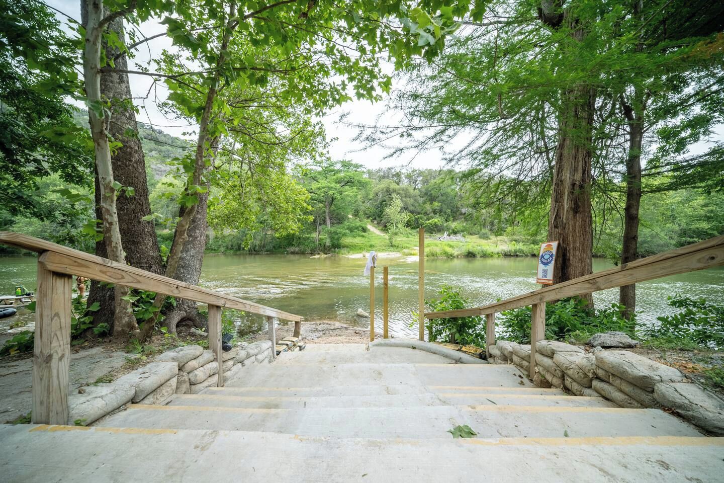 A wooden staircase leading down to a river, surrounded by trees and a stone wall.