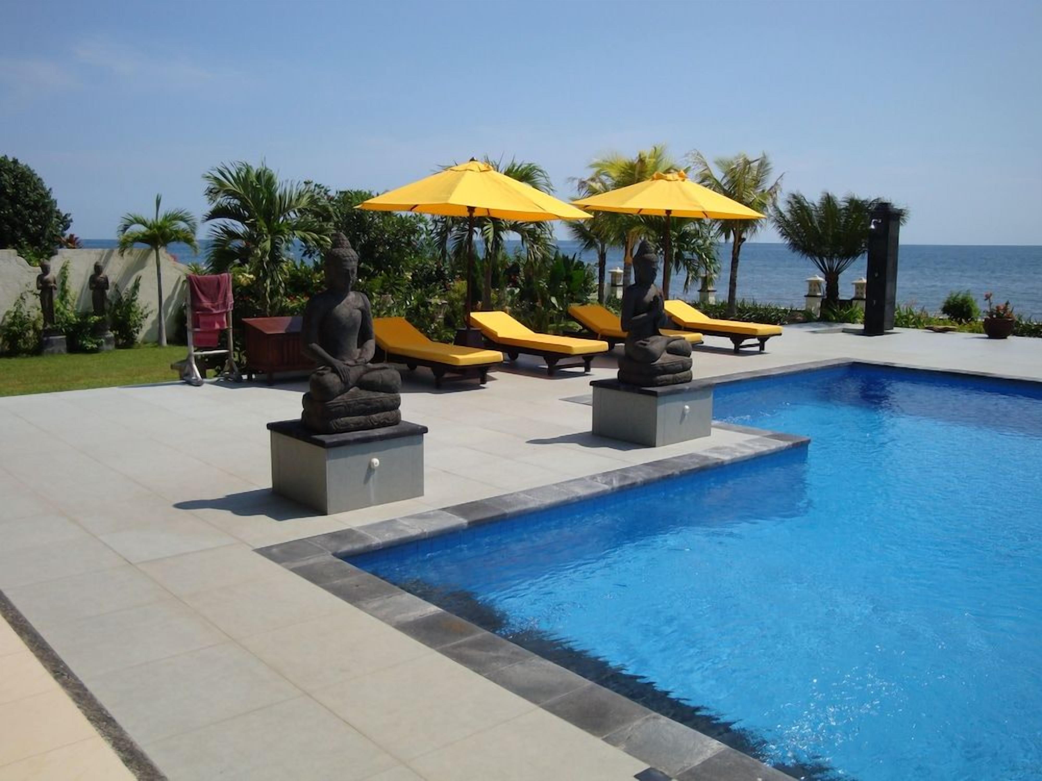 A swimming pool with sun loungers and umbrellas, surrounded by palm trees and a view of the sea.
