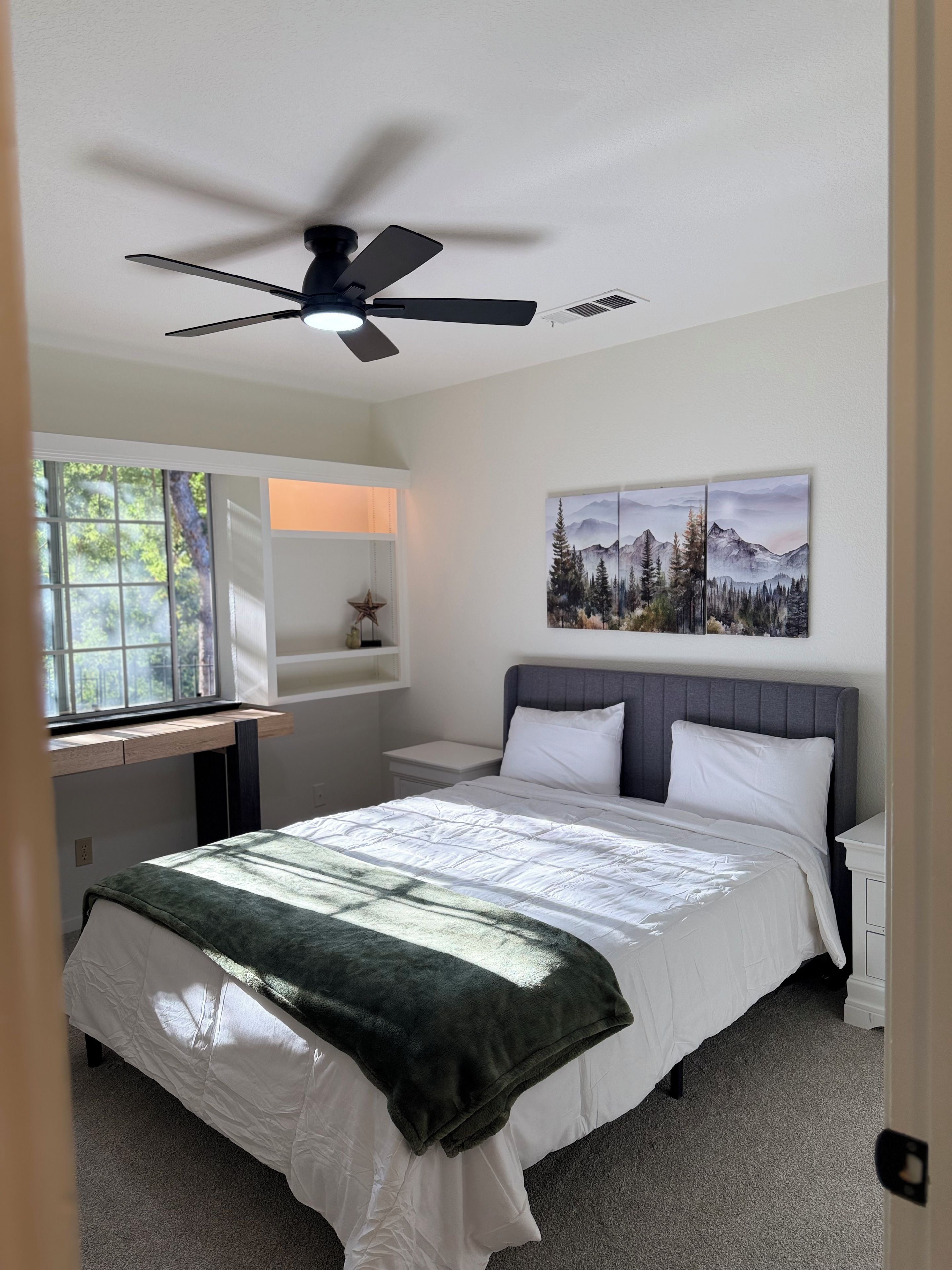 A bedroom with a bed, a ceiling fan, a window, a desk, and a wall-mounted painting.