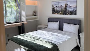 A bedroom with a bed, a ceiling fan, a window, a desk, and a wall-mounted painting.
