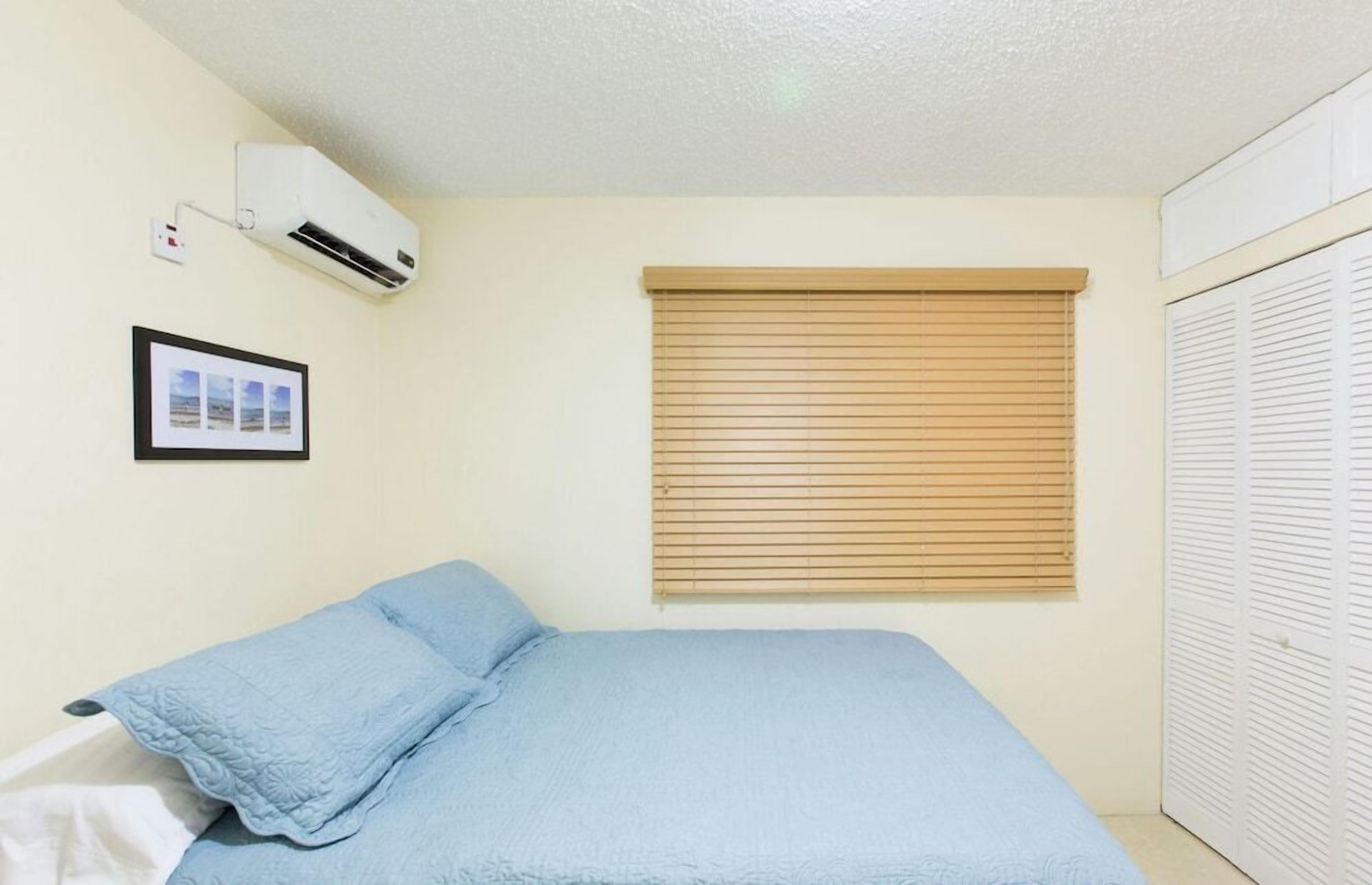 A bedroom with a bed, an air conditioning unit, a window with blinds, a framed picture on the wall, and a white wardrobe.