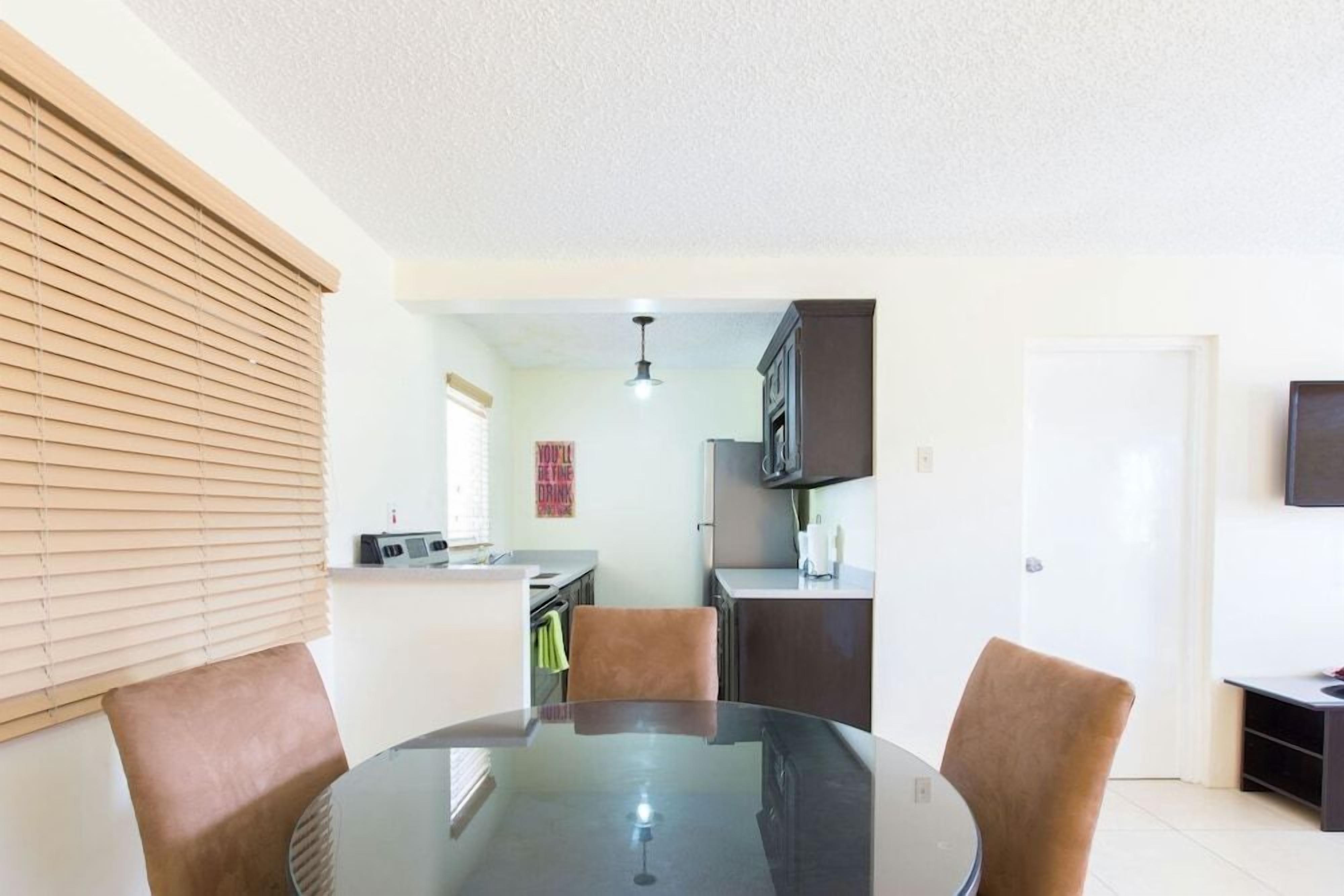 A dining area with a round table and four chairs, a kitchen in the background, and a white door.