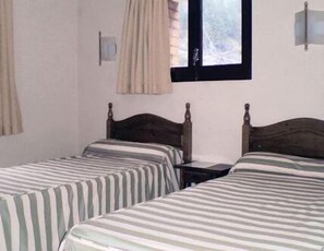 Una habitación con dos camas individuales, una mesita, una ventana con cortinas y una lámpara fijada a la pared.
