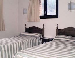 Una habitación con dos camas individuales, una mesita, una ventana con cortinas y una lámpara fijada a la pared.