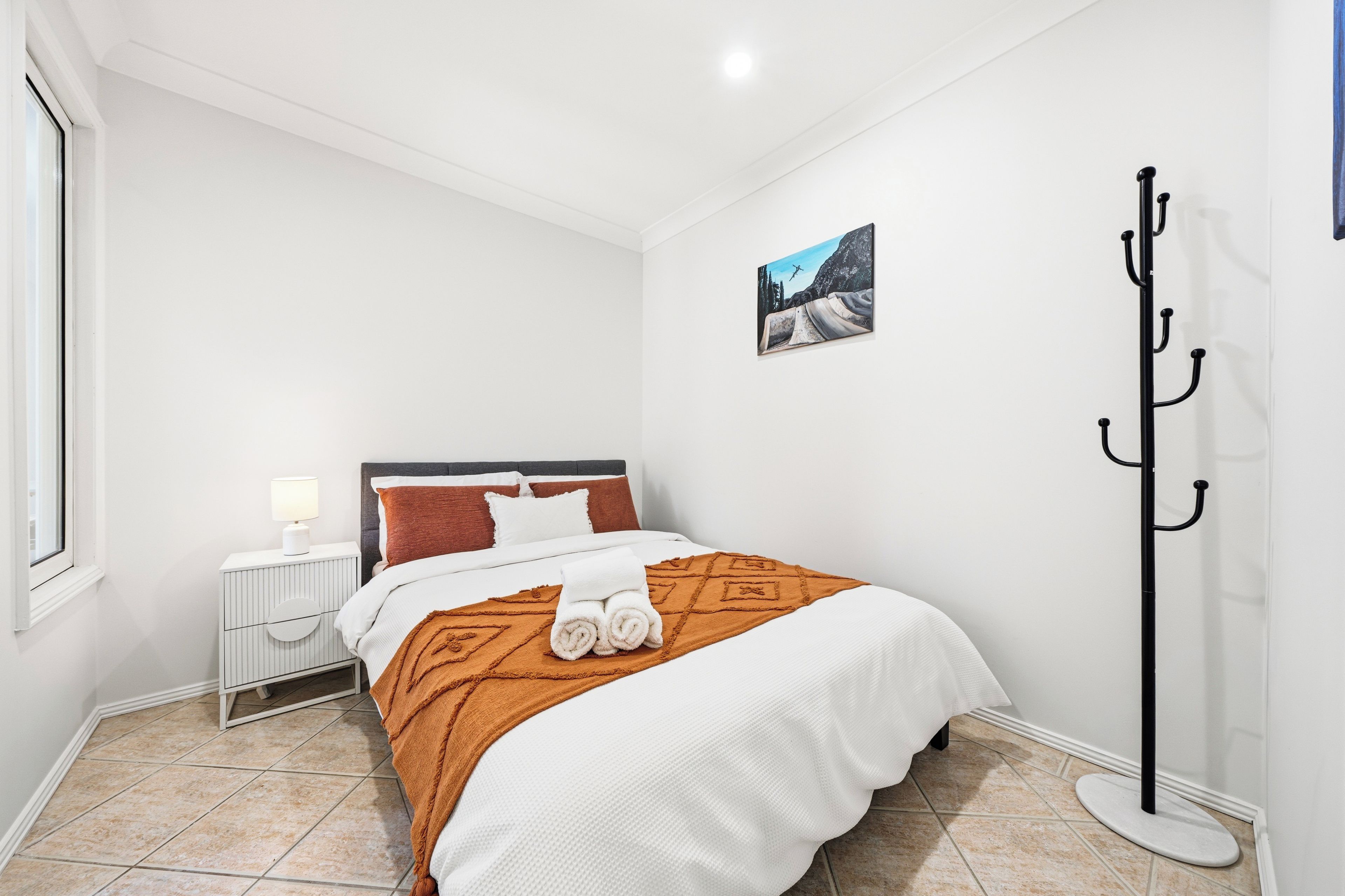 Une chambre d’hôtel avec un lit, une table de chevet, une lampe, un tableau et un porte-manteau.