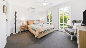 Un dormitorio con cama, mesitas de noche, una silla, un escritorio, un ventilador de techo y una ventana con vistas a zonas verdes.