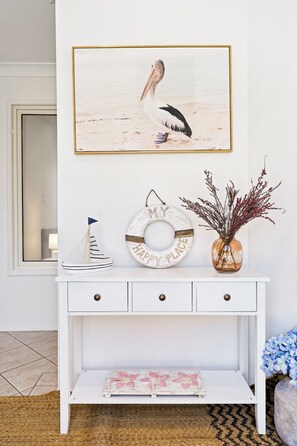 A white console table with a framed seascape painting, a decorative plate, and a vase with dried plants.
