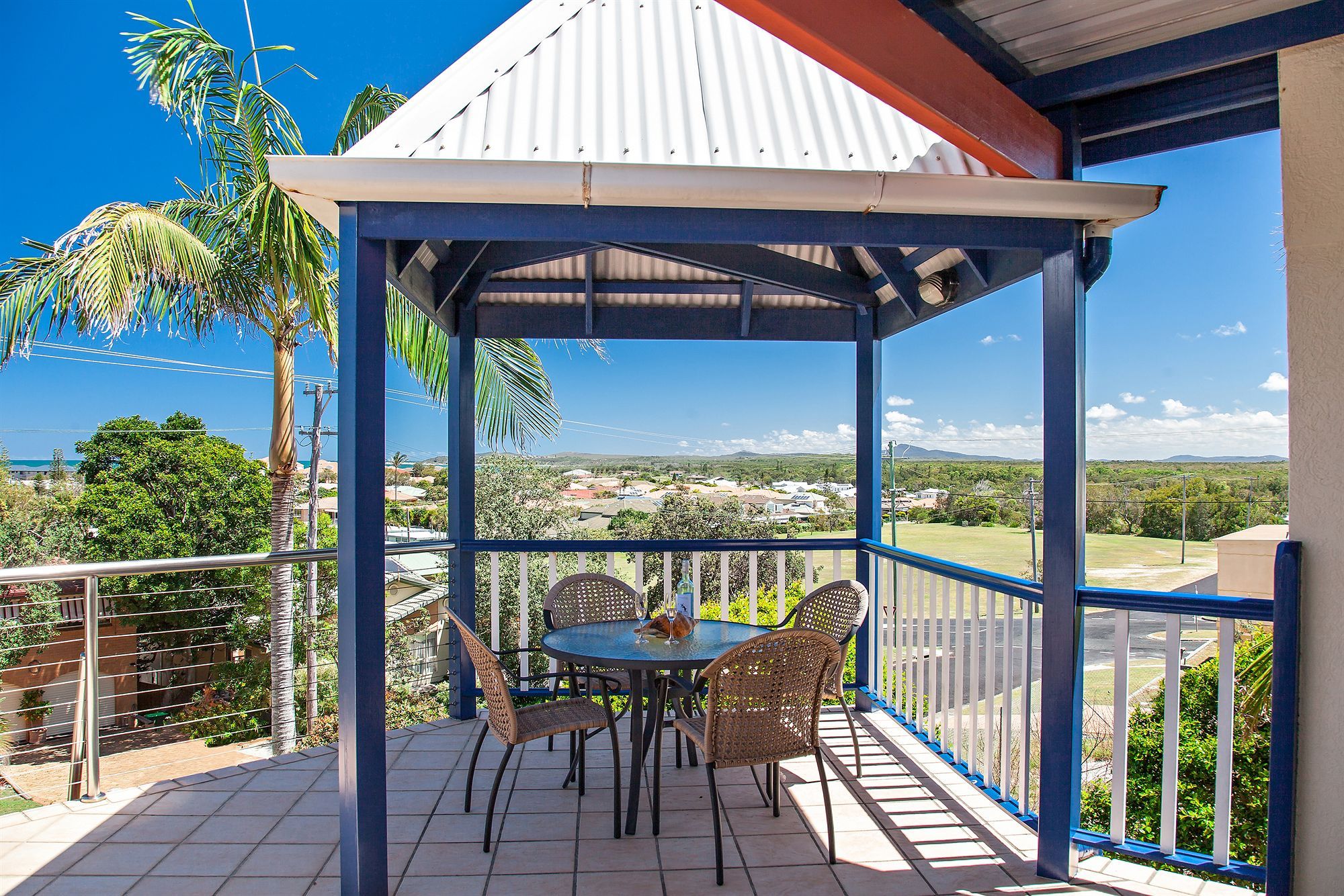 A covered outdoor seating area with a table and chairs, overlooking a residential area with houses and greenery.