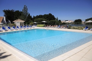 Une piscine rectangulaire à l’eau d’un bleu limpide, entourée de chaises longues et de parasols, avec en arrière-plan une vue sur des arbres et des bâtiments.