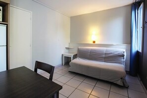 Una habitación pequeña con una cama, una mesa y una silla. Incluye una luz en la pared y una puerta.