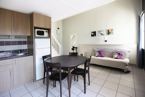 A compact living space with a kitchenette, a dining area with a round table and chairs, a sofa with cushions, and a staircase leading to another level. - Belambra Clubs Les Grands Espaces (Saint-Jean-de-Monts)
