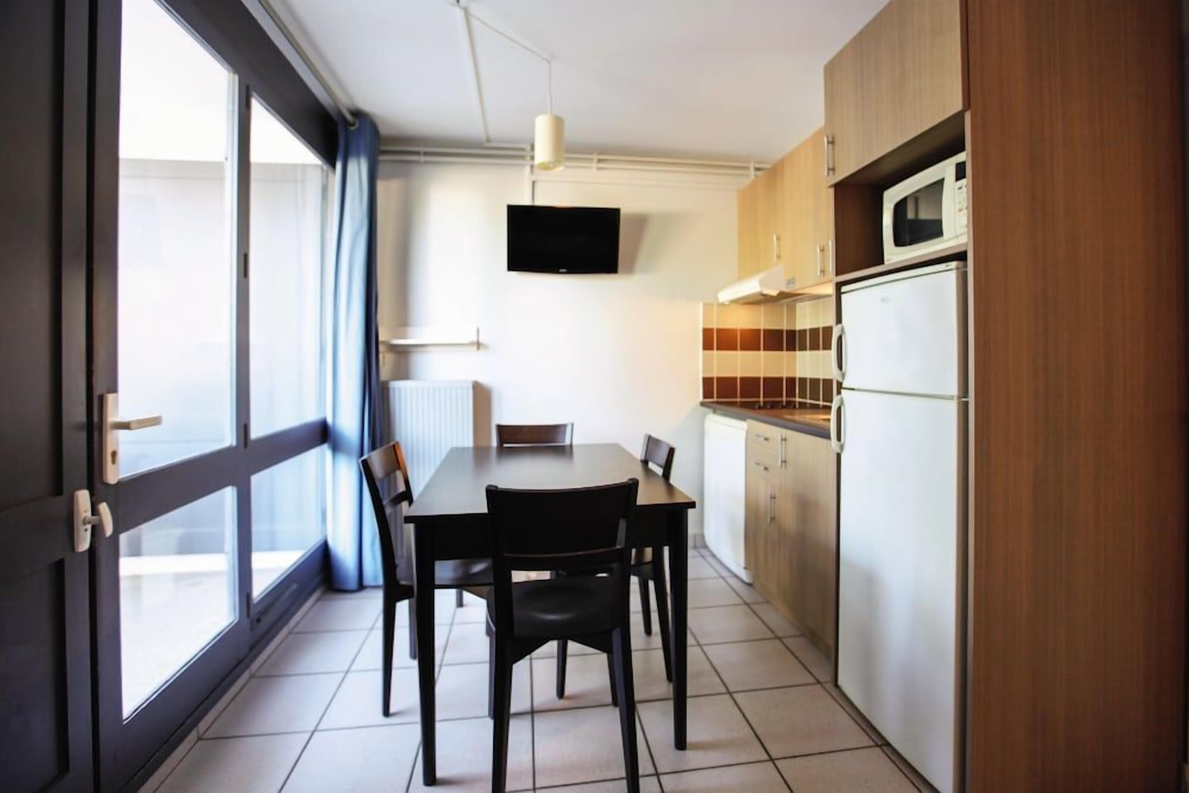 A compact kitchen with a dining table, chairs, a TV mounted above the counter, and a refrigerator.