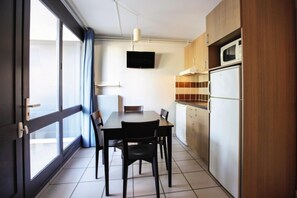 A compact kitchen with a dining table, chairs, a TV mounted above the counter, and a refrigerator. - Belambra Clubs Les Grands Espaces (Saint-Jean-de-Monts)