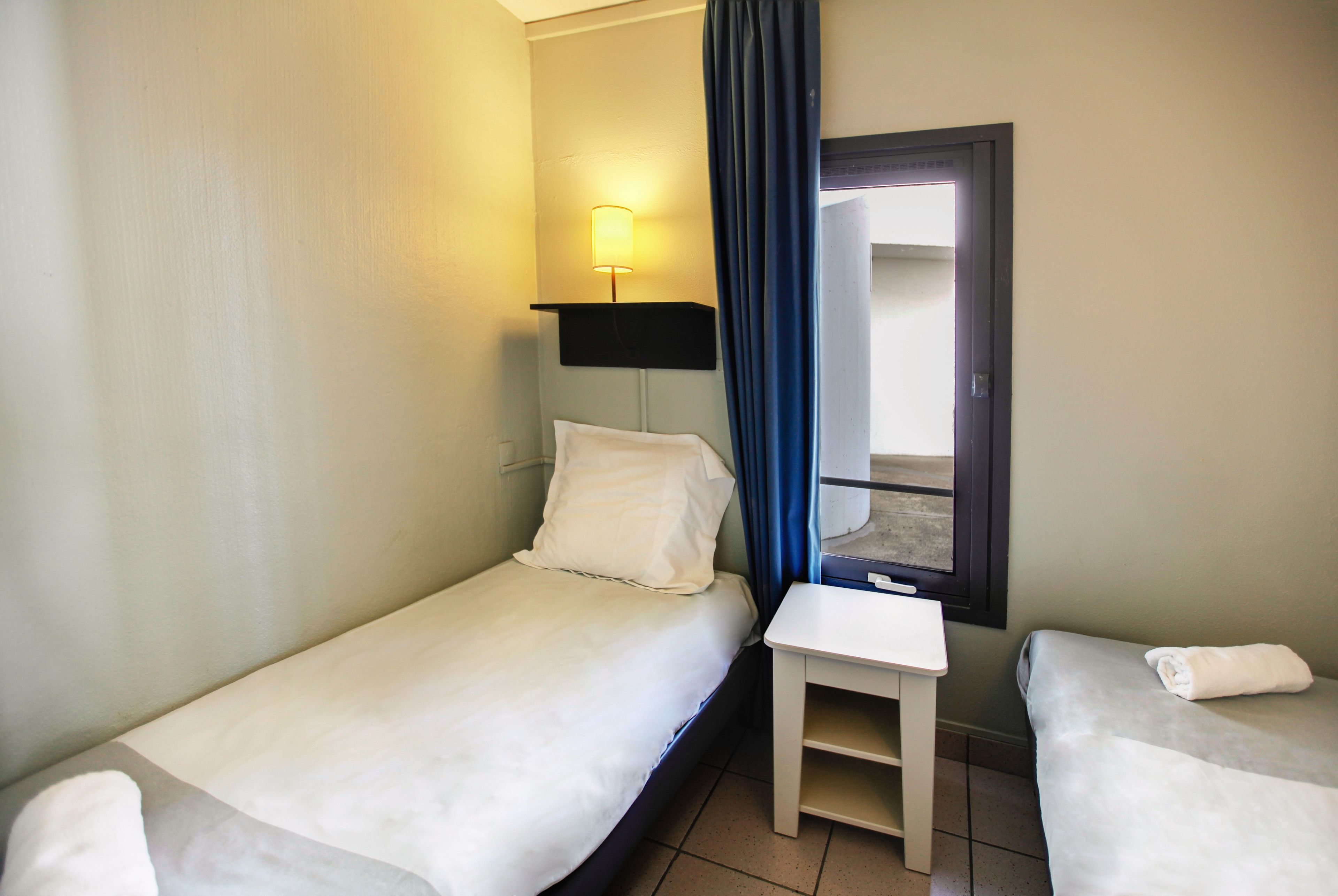 A small hotel room with two single beds, a nightstand, a lamp, and a window with curtains.